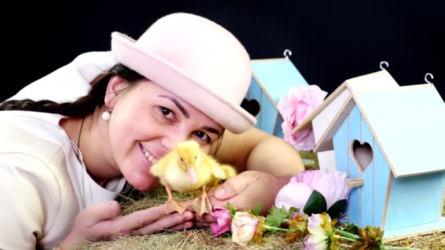 Portrait,-a-pretty-young-woman-with-two-pigtails-and-in-a-funny-pink-hat-playing-with-small-yellow-ducklings.-Studio-video-with-thematic-decor