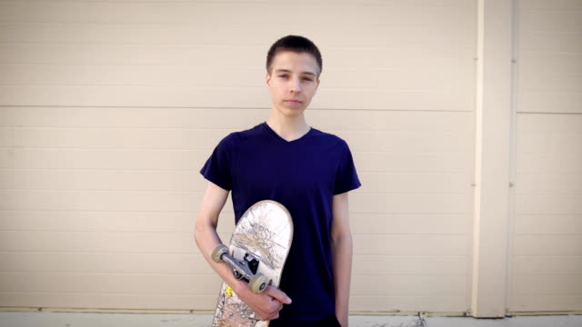 Image-of-handsome-teenager-standing-in-the-street-and-holding-skateboard-in-his-hands.-Boy-spending-free-time-in-skateboarding-park-demonstrating-healthy-lifestyle-and-looking-at-the-camera