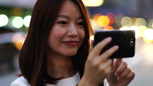 Mujer-asiática-atractiva-usando-el-teléfono-en-la-ciudad.-Ojos-oscuros-y-cabello-oscuro.-Fondo-lleno-de-gente-y-urbano.