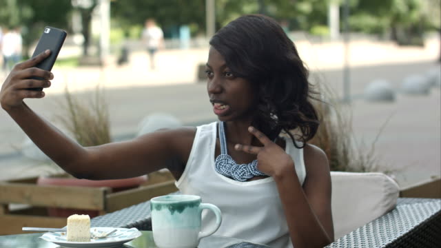 Attraktive-junge-afrikanische-Frau-Lächeln-auf-den-Lippen-und-ein-Selbstporträt-mit-ihrem-Smartphone-dabei-allein-im-Café-genießen-Sie-eine-Mahlzeit-im-Freien-sitzen