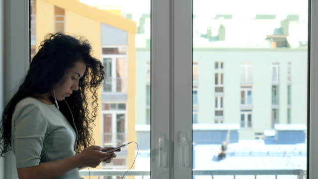 Curly-brunette-messaging-with-someone-listening-a-music-near-the-window