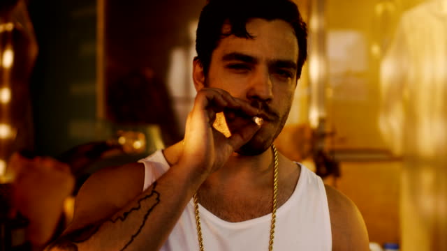 Smoking-Brutal-Gang-Member-with-Gold-Chain-Looks-into-Camera-with-Defiance.-Underground-Drug-Laboratory-is-in-Background.