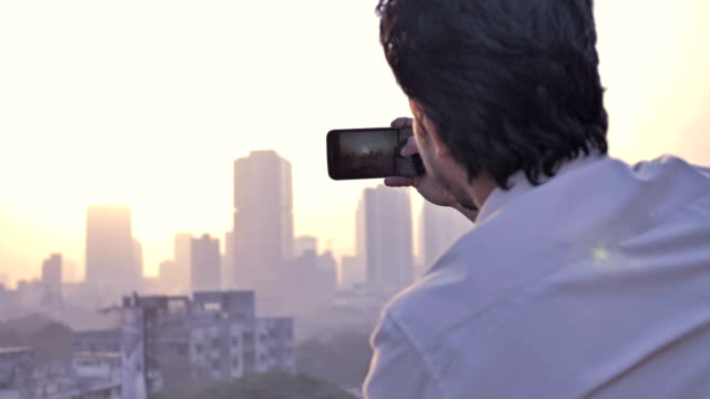 Un-hombre-joven-clic-en-fotos-de-una-puesta-de-sol-sobre-el-horizonte-de-una-ciudad-usando-un-teléfono-celular