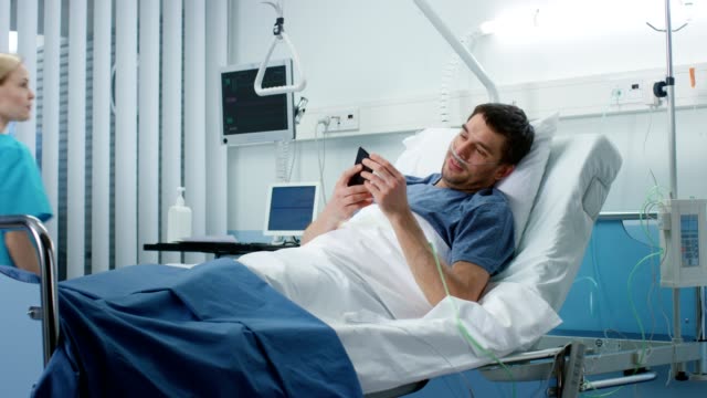 Recovering-Patient-Uses-Smartphone-and-Smiles-while-Lying-on-a-Bed-in-the-Hospital.-Friendly-Nurse-Comes-in-to-Check-on-Him.