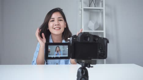 Glücklich-lächelnd-asiatische-Frau-oder-Beauty-Blogger-mit-Pinsel-und-Kamera-Videoaufnahme-und-winkenden-Hand-zu-Hause.-Videoblog-Blogging-Menschen-Schönheitskonzept.-Dolly-erschossen.