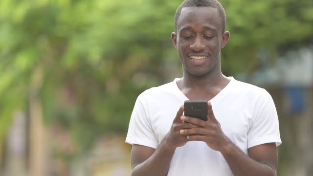 Young-happy-African-man-smiling-while-using-phone-in-the-streets-outdoors