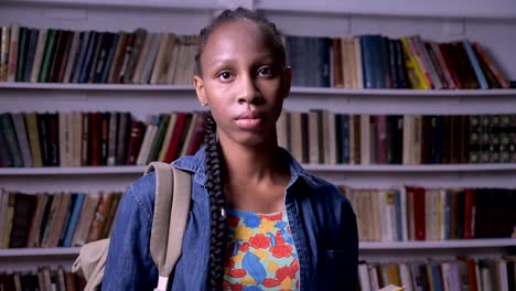 Young-african-american-woman-with-backpack-reading-book-in-library-and-looking-at-camera,-serious-and-thoughtful,-bookshelves-background