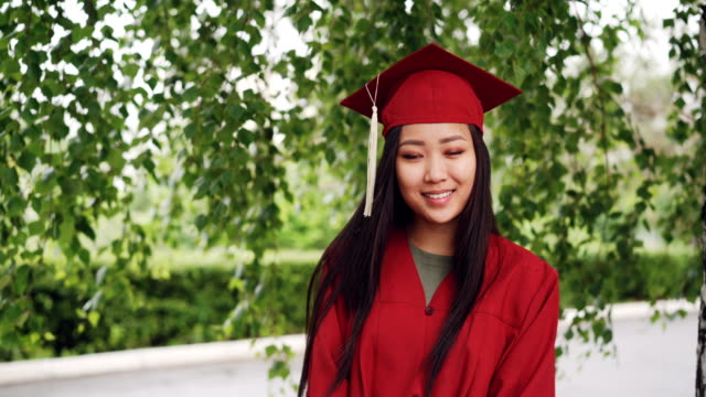 Portrait-of-attractive-Asian-girl-successful-graduating-student-in-gown-and-mortar-board-standing-on-campus,-smiling-and-looking-at-camera.-Youth-and-education-concept.