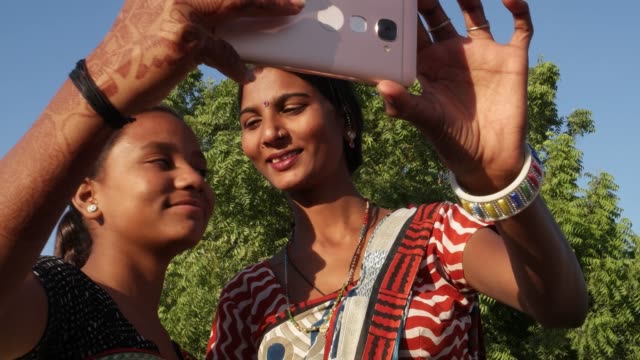 Dos-mujeres-riendo-sonriendo-tomando-selfie-video-foto-junto-con-un-teléfono-inteligente-ayuda-mutua-vinculación-amor-mano-ayudar-a-tiro-medio-en-traje-tradicional-de-Rajasthani-en-soleado-entorno-al-aire-libre