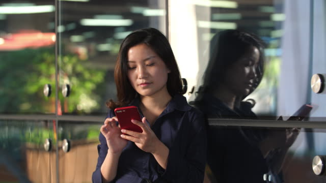 Joven-empresaria-asiática-tocando-la-pantalla-del-teléfono-móvil-que-se-inclina-el-vidrio-de-ventana-de-la-oficina,-4k