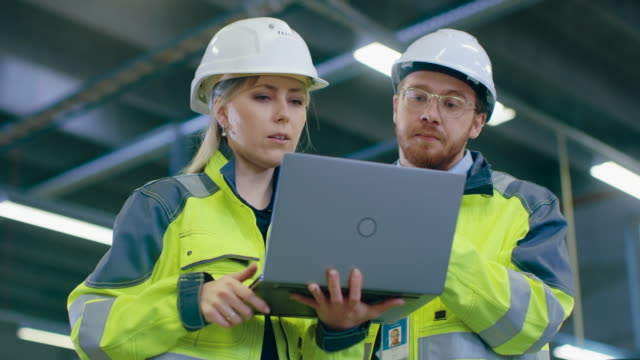 Low-Angle-Shot-of-Male-and-Female-Industrial-Engineers-Work-on-a-Manufacturing-Plant,-They-Discuss-Project-while-Using-Laptop.In-the-Background-Industrial-Manufacturing-Plant.