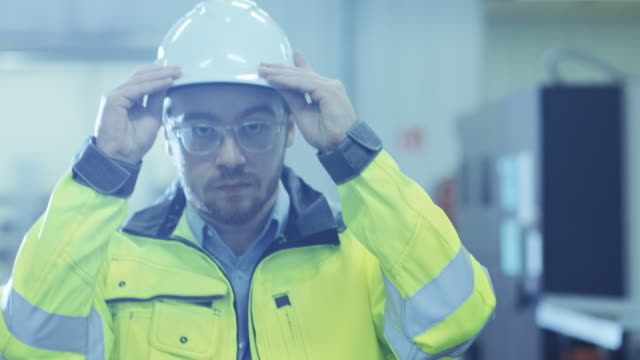 Industrial-Engineer-Wearing-Protective-Clothing-Puts-on-Hard-Hat-and-Walks-Through-Modern-Manufacturing-Facility-with-Automatic-Machinery-Working-in-Background.