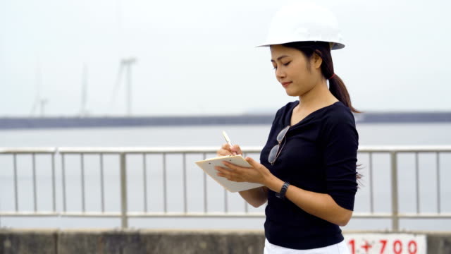 Asian-female-engineer-checks-job-The-background-is-a-large-reservoir-and-rotating-wind-turbines.