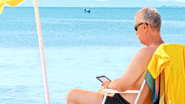 Hombre-maduro-en-la-playa-leyendo-el-lector-electrónico