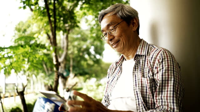 Asiatischer-senior-Mann-mit-Tablet,-allein-auf-der-Bank-im-Park-sitzen