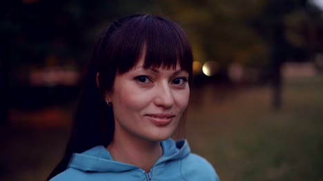 Close-up-portrait-of-beautiful-brunette-with-ponytail-wearing-bright-hoody-looking-at-camera-and-smiling-standing-in-the-park-in-the-evening-in-autumn.