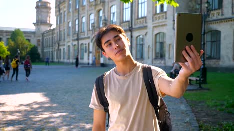 Glad-asian-male-is-taking-selfies-on-his-tablet-and-standing-straight,-near-school-building,-separated,-summertime