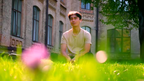 Calm-nice-asian-man-in-white-earphones,-concentrated-on-music-listening-and-having-rest-while-sitting-on-the-grass-in-park
