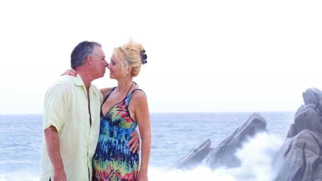 An-older-couple-kiss-each-other-on-the-beach-and-then-smile,-with-waves-crashing-on-rocks-behind-them