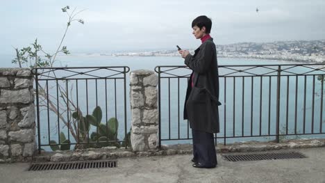 Mujer-enviando-mensajes-de-texto-sms-usando-la-aplicación-de-teléfono-inteligente-en-la-ciudad-con-vista-a-la-costa