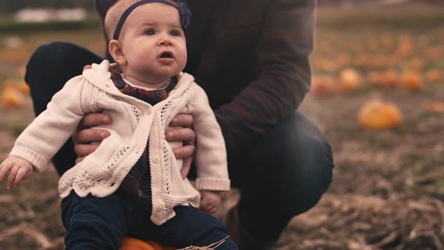 Close-up-of-a-baby-sitting-on-a-pumpkin-at-a-pumpkin-patch,-with-her-dad-holding-her-up
