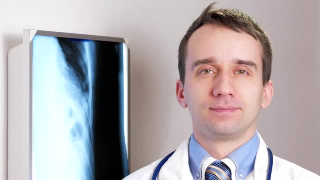 A-young-doctor-looks-at-the-camera-and-smiles.-Against-the-background-hanging-x-ray-of-the-patient.-Shirt-with-a-tie-and-a-stethoscope-on-the-neck