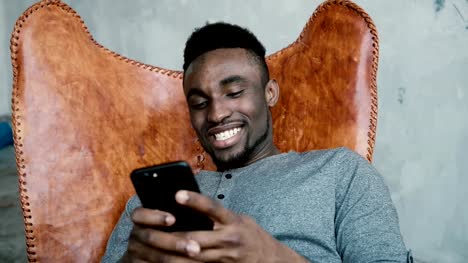 Portrait-des-afrikanischen-Mann-sitzt-im-Stuhl,-mit-Smartphone.-Mann-liest-Nachrichten-und-lacht.-Guy-Eingabe-Massagen,-Lächeln