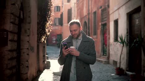 Young-smiling-man-walking-in-city-at-morning-and-using-smartphone.-Guy-browse-the-Internet,-texting-with-someone