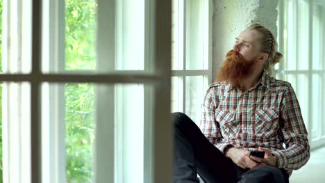 Sad-bearded-man-sms-texting-using-smartphone-sit-at-windowsill-at-home