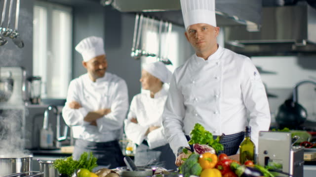 Famous-Chef-of-a-Big-Restaurant-Prepares-Dishes-and-Smiles-On-Camera.-In-the-Background-Two-Apprentices-and-Modern-Kitchen.
