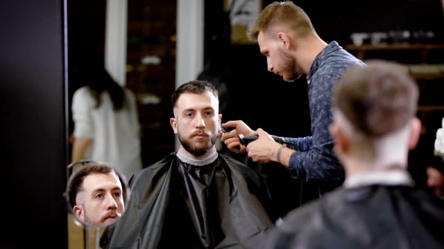 Stylish-barber-is-shaving-temples-with-hairclipper-of-a-bearded-man-sitting-on-armchair-in-the-barbershop-and-looking-at-the-mirror.-Young-coiffeur-is-cutting-the-hair-of-his-client-in-the-black-cape