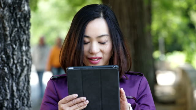 Asiático-joven-mujer-utilizando-Digital-Tablet-y-sonriente-al-aire-libre