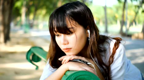 Hermosa-joven-inconformista-triste-mujer-asiática-emocionalmente-escuchar-música-en-auriculares-smartphone,-sentado-en-el-Banco-del-parque-en-luz-del-sol-del-atardecer-de-verano.
