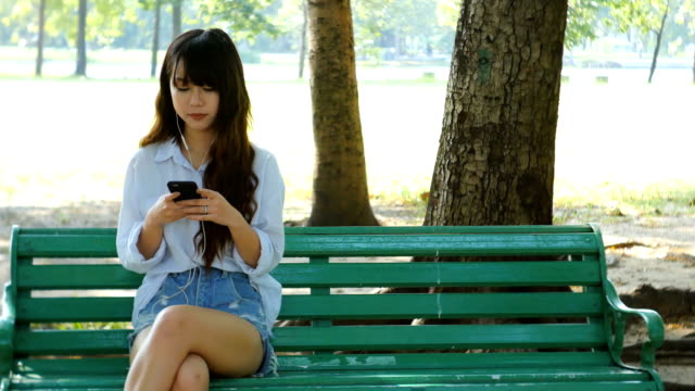 Nette-Frau-ist-lesen-angenehm-SMS-auf-Handy-beim-Sitzen-im-Park-in-warmen-Frühlingstag,-wunderschöne-Weibchen-ist-Musik-im-Kopfhörer-hören-und-suchen-Informationen-über-Zelle-Telefon