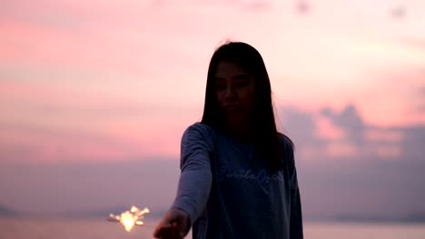 Junge-asiatische-Frau-steht-mit-Wunderkerze-am-Strand-bei-Sonnenuntergang-in-Zeitlupe.