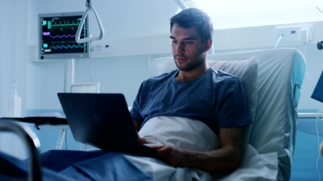 In-the-Hospital,-Recovering-Male-Patient-Uses-Laptop-while-Lying-on-the-Bed.-Working-even-when-Sick-and-in-Hospital.-Nurse-Checks-Drop-Counter.