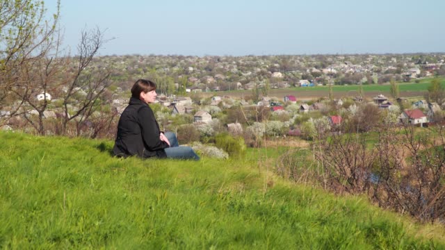 Eine-junge-Frau-sitzt-auf-einem-Hügel-und-schaut-auf-das-Haus