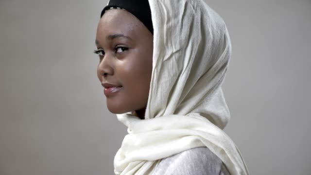Young-african-muslim-girl-in-hijab-is-turning-head-and-watching-at-camera,-smiling,-religioun-concept,-grey-background