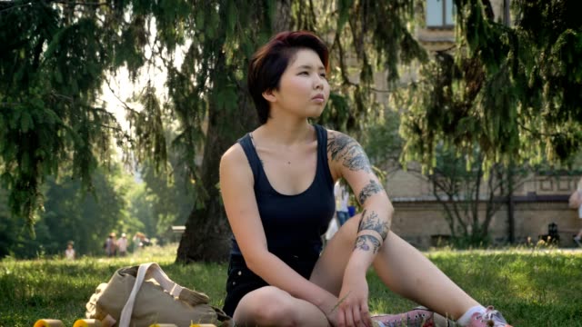 Trendy-young-asian-female-hipster-with-tattoo-sitting-on-grass-in-park-near-university,-thoughtful