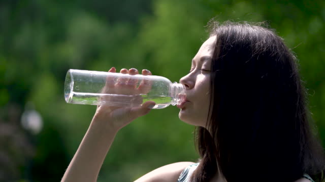 Close-up-of-a-happy-looking-asian-woman-drinks-water-from-a-transparent-bottle.-Sunrays-are-on-her-face.