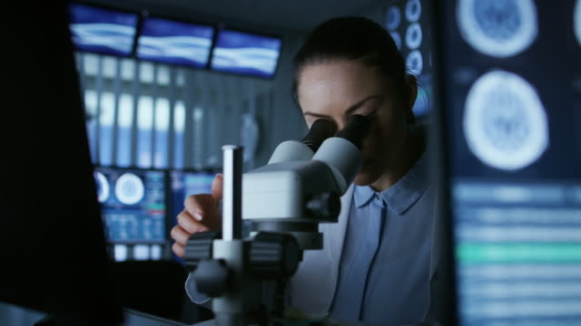 Weibliche-medizinische-Forschungswissenschaftler-Blick-durch-das-Mikroskop-Arten-erfassten-Daten-in-den-Computer.-Labor.-Im-Labor-mit-mehreren-Bildschirmen-zeigen,-MRI-/-CT-Gehirn-Bilder-scannen.