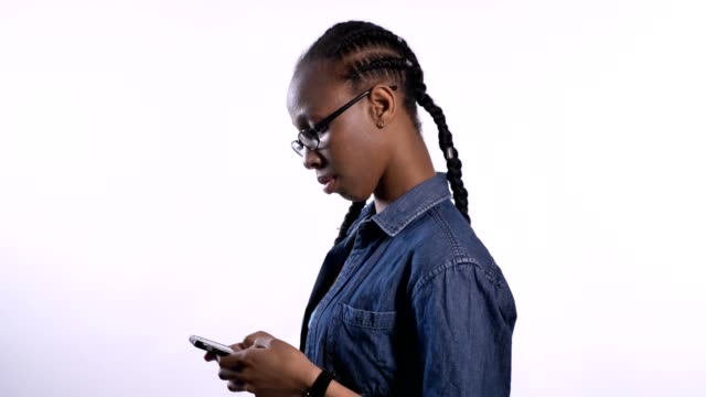 Young-beautiful-african-american-woman-in-glasses-texting-on-phone-and-looking-at-camera,-profile-of-black-model-in-studio-with-phone,-serious