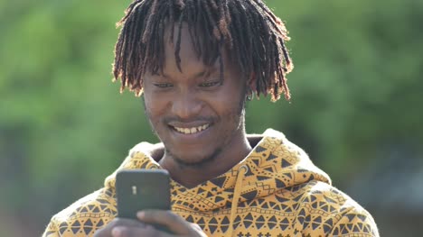 Feliz-guapo-africano-joven-sonriendo-mientras-usa-el-teléfono-en-las-calles-al-aire-libre