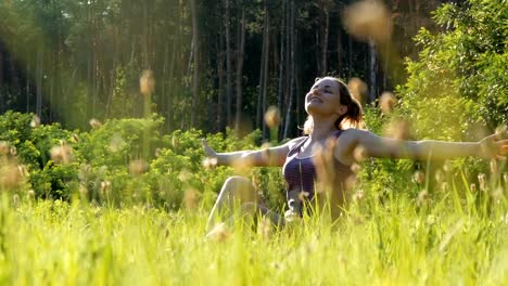 Fröhliche-Frau-sitzt-auf-dem-grünen-Rasen-hebt-gerne-ihre-Hände-zum-Himmel-auf-malerischen-Feld-bei-Sonnenuntergang-Hintergrund