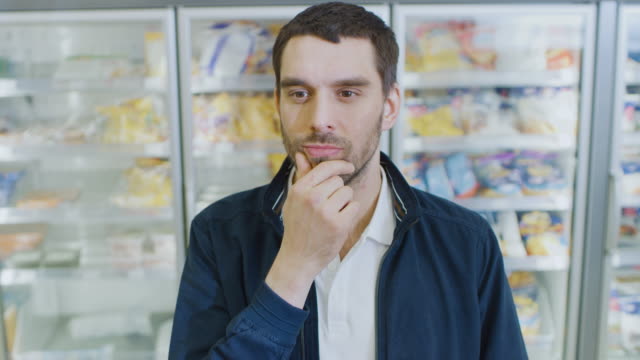 Portrait-Shot-of-the-Handsome-Man-Thinking-Very-Hard-and-Choosing-Tin-Can-from-the-Canned-Goods-Section,-Plaves-it-i-His-Shopping-Cart.-In-the-Background-Frozen-Goods-Section-of-the-Store.