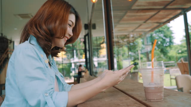 Cheerful-happy-asian-young-woman-sitting-drinking-iced-green-tea-in-cafe-using-smartphone-for-talking,-reading-and-texting.-Women-lifestyle-concept.