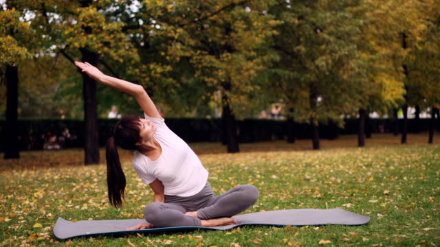 Beautiful-girl-is-concentrated-on-yoga-practice-sitting-on-mat-in-park-in-lotus-position-and-bending-sideways-raising-arm.-Healthy-lifestyle-and-recreation-concept.