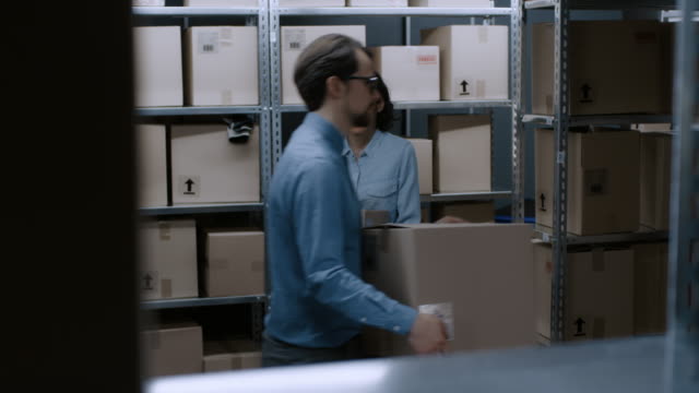 Female-Warehouse-Worker-Puts-Cardboard-Box-of-a-Shelf-and-Smiles-Charmingly.-In-the-Background-Rows-of-Shelves-Full-of-Cardboard-Boxes-and-Parcels-Filled-with-Products-Ready-for-Shipment.