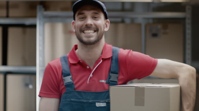Portrait-of-Handsome-Warehouse-Worker-in-Uniform-Holds-Cardboard-Box-Package-and-Smiles.