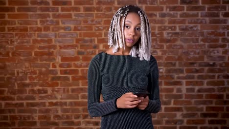 Young-African-girl-with-dreadlocks-using-a-smartphone-looking-at-the-camera-and-smiling,-Brick-wall-in-the-background.
